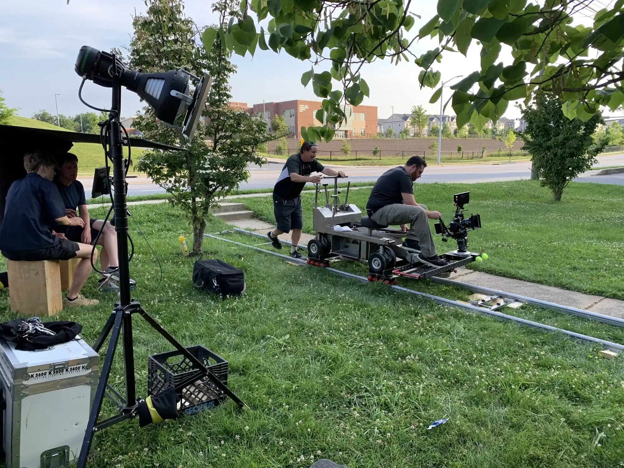 Camera operator on dolly track filming alongside lighting setup outdoors