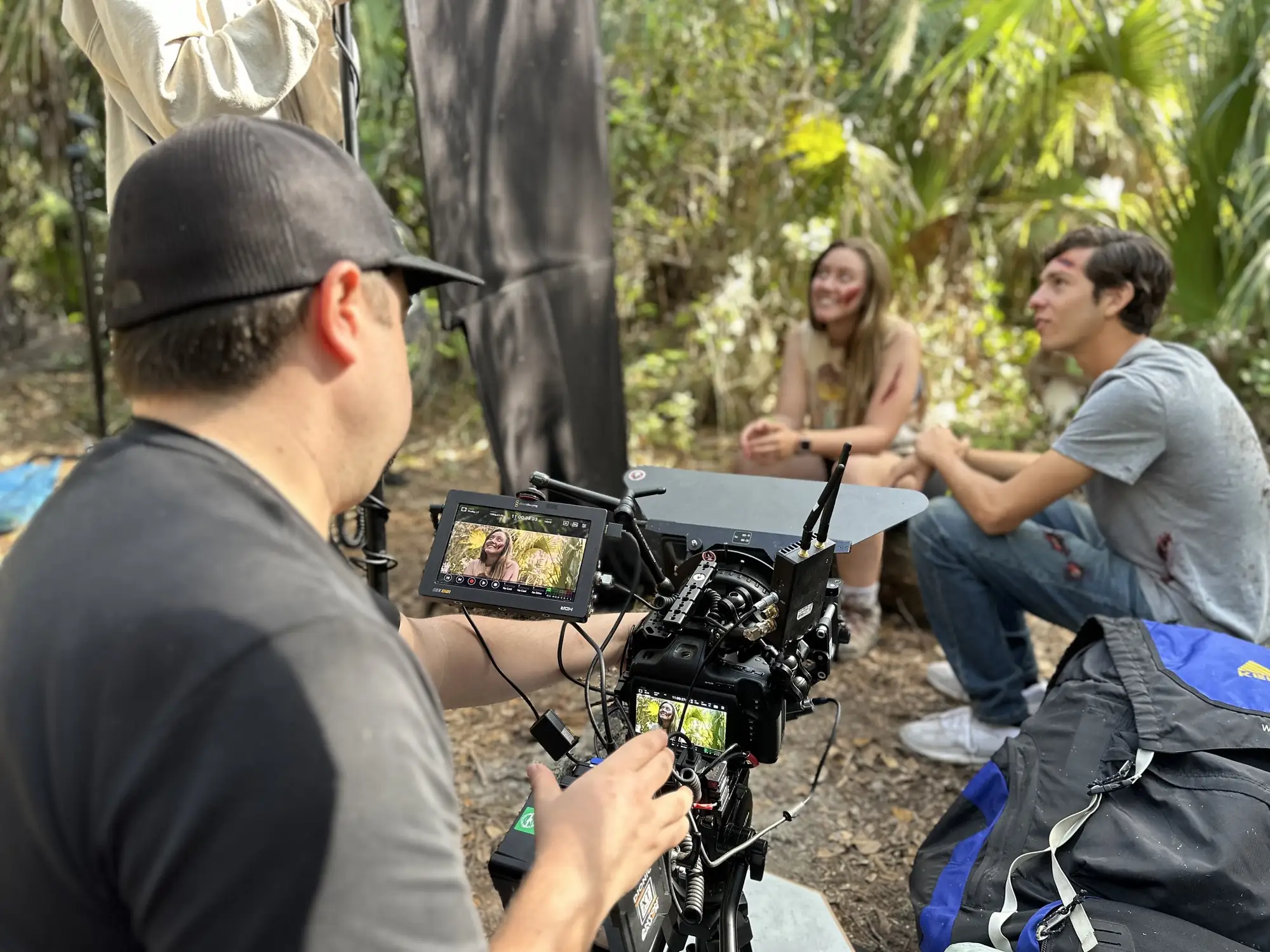 Camera operator filming two actors sitting in a palmetto forest